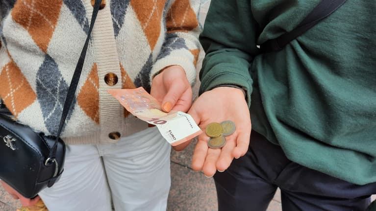 Kaksi ihmistä pitää kädessään rahaa. Toisella on 10 euron seteli ja toisella on kolikoina 4,50.